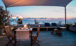 Cozy outdoor dining area with wooden furniture, a table set for two, and a stunning sunset view over the ocean. The scene features soft lighting from candles and a stylish canopy, creating a romantic atmosphere.