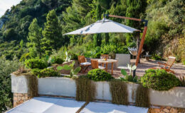 Outdoor terrace with wooden furniture, a large umbrella, and lush greenery overlooking a scenic hillside. The space features potted plants and a tranquil atmosphere, ideal for relaxation and outdoor dining.