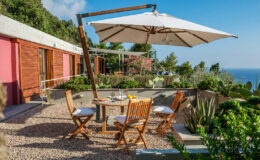 Outdoor dining area with wooden furniture and a large umbrella, surrounded by lush greenery and a scenic ocean view. Perfect for relaxing and enjoying meals in a tranquil setting.