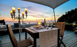 Romantic outdoor dining setup at sunset, featuring a candlelit table with elegant chairs, a bottle of wine, and a scenic view of the ocean.