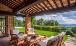 Cozy outdoor seating area with wicker chairs and a coffee table, overlooking a lush green landscape and a swimming pool under a cloudy sky. Perfect for relaxation and enjoying nature.