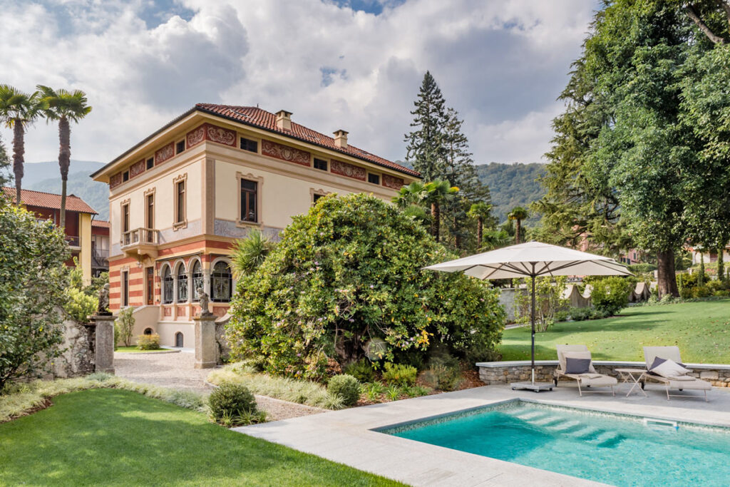 Elegant villa with a swimming pool surrounded by lush greenery and landscaped gardens, featuring a patio area with sun loungers and an umbrella, set against a backdrop of trees and a cloudy sky. | Only on SopranoVillas