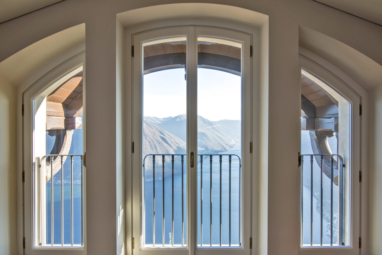 Elegant arched windows with a view of mountains and a serene lake, showcasing a modern architectural design and natural light. | Only on SopranoVillas