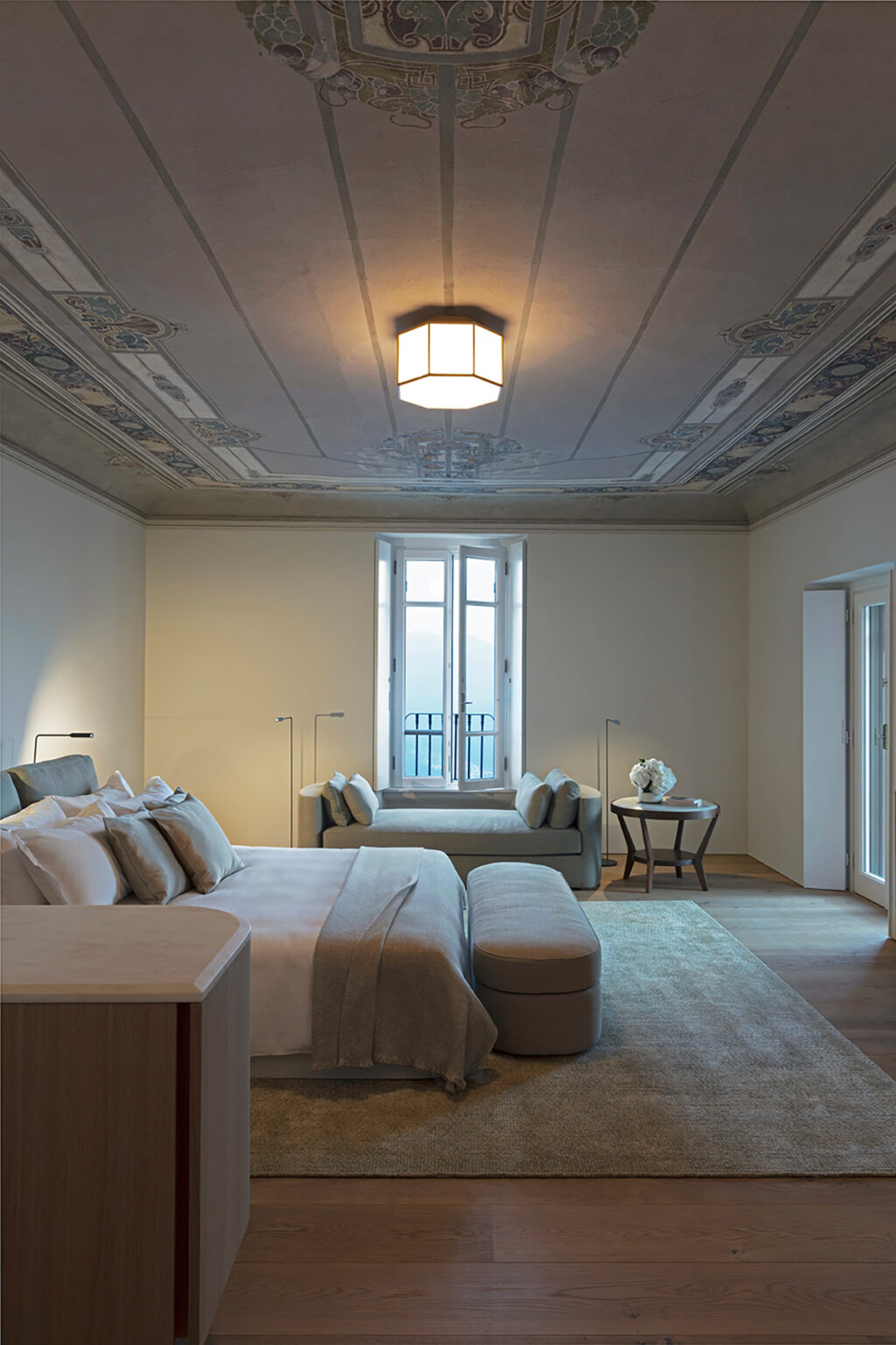 Spacious modern bedroom featuring a comfortable bed with decorative pillows, a cozy seating area, large windows allowing natural light, and elegant ceiling details. | Only on SopranoVillas