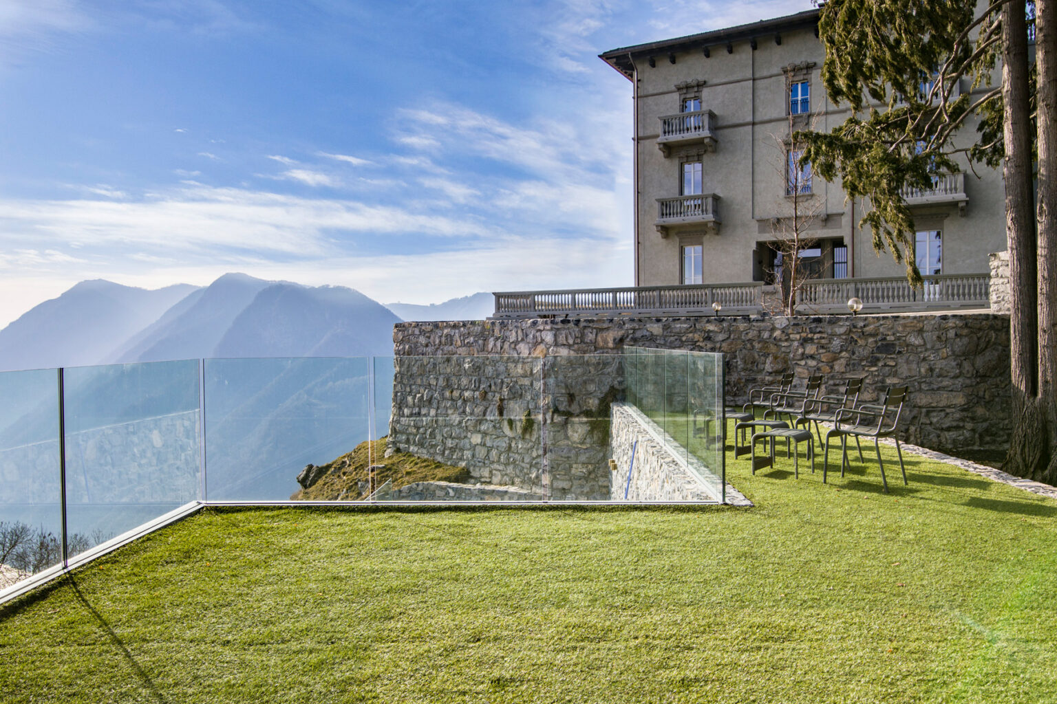 Modern glass railing overlooking a scenic mountain view, with a well-maintained lawn and a historic stone building in the background. Ideal for architectural and landscape photography. | Only on SopranoVillas