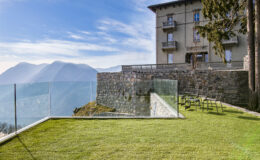 Modern glass railing overlooking a scenic mountain view, with a well-maintained lawn and a historic stone building in the background. Ideal for architectural and landscape photography.
