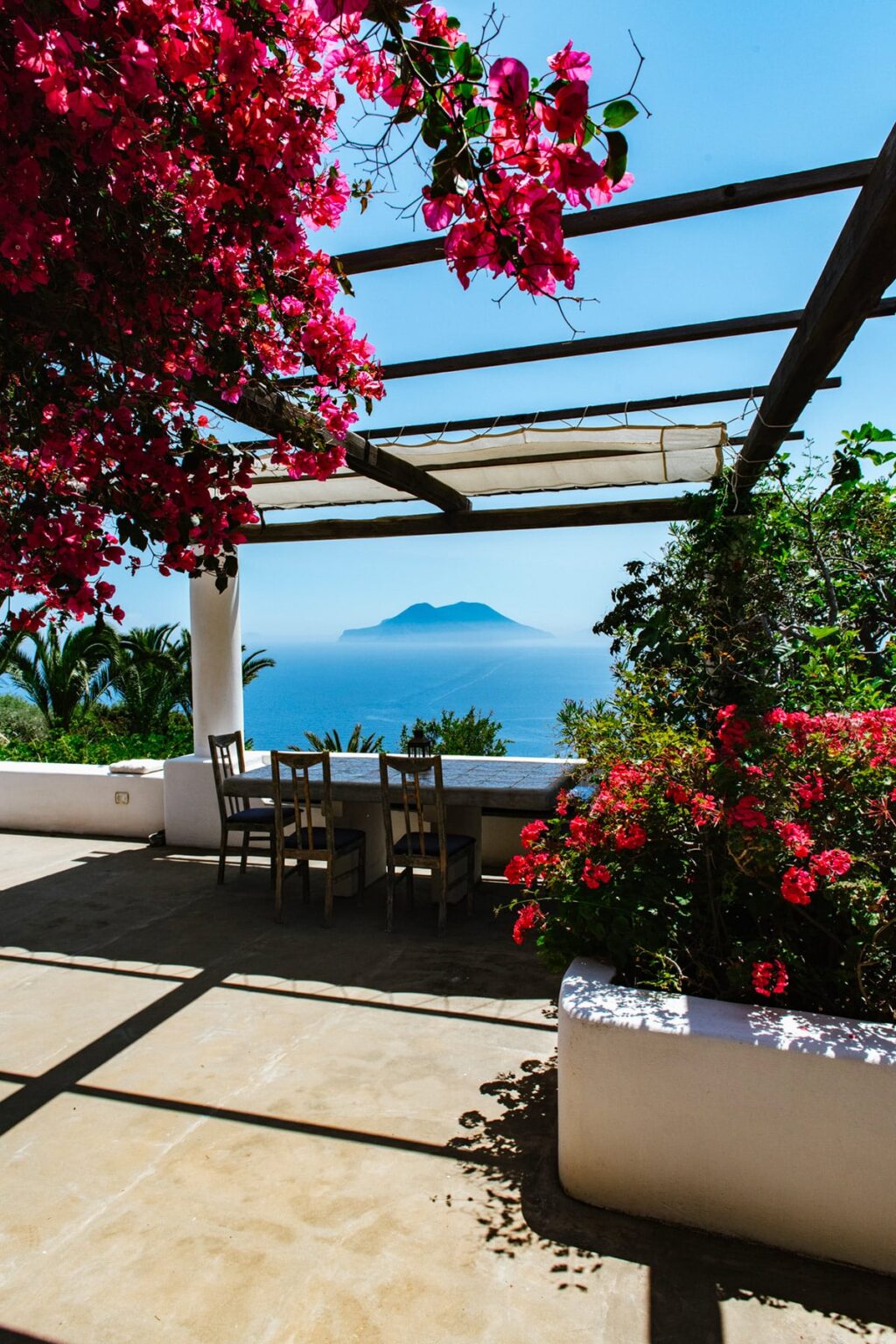 Scenic outdoor dining area with a view of a distant island, surrounded by vibrant bougainvillea flowers and lush greenery, ideal for relaxation and enjoying nature. | Only on SopranoVillas