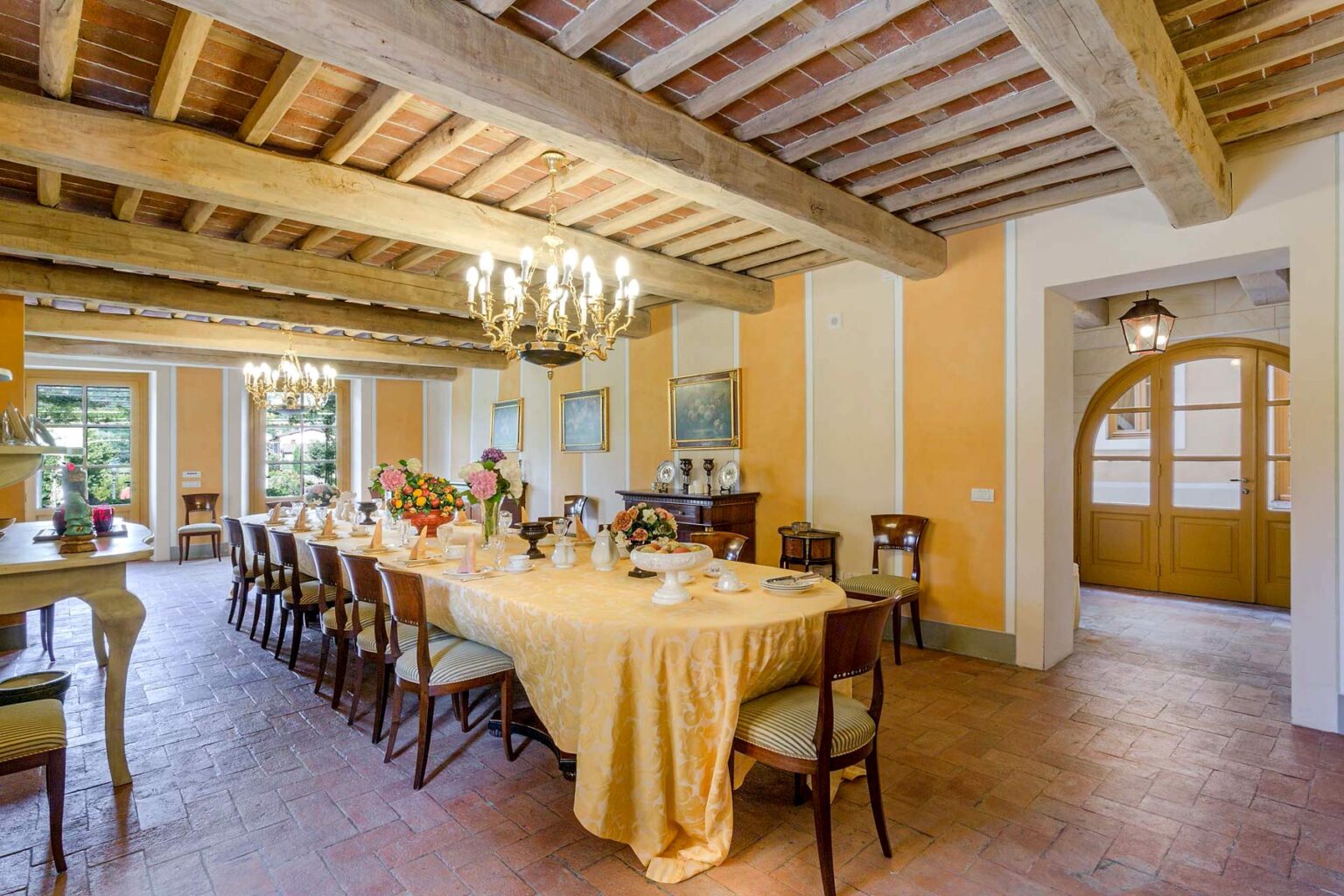 Spacious dining room with a long table set for a meal, featuring a yellow tablecloth, wooden beams on the ceiling, and warm orange walls, illuminated by a chandelier. Large windows provide natural light and a view of greenery outside. | Only on SopranoVillas