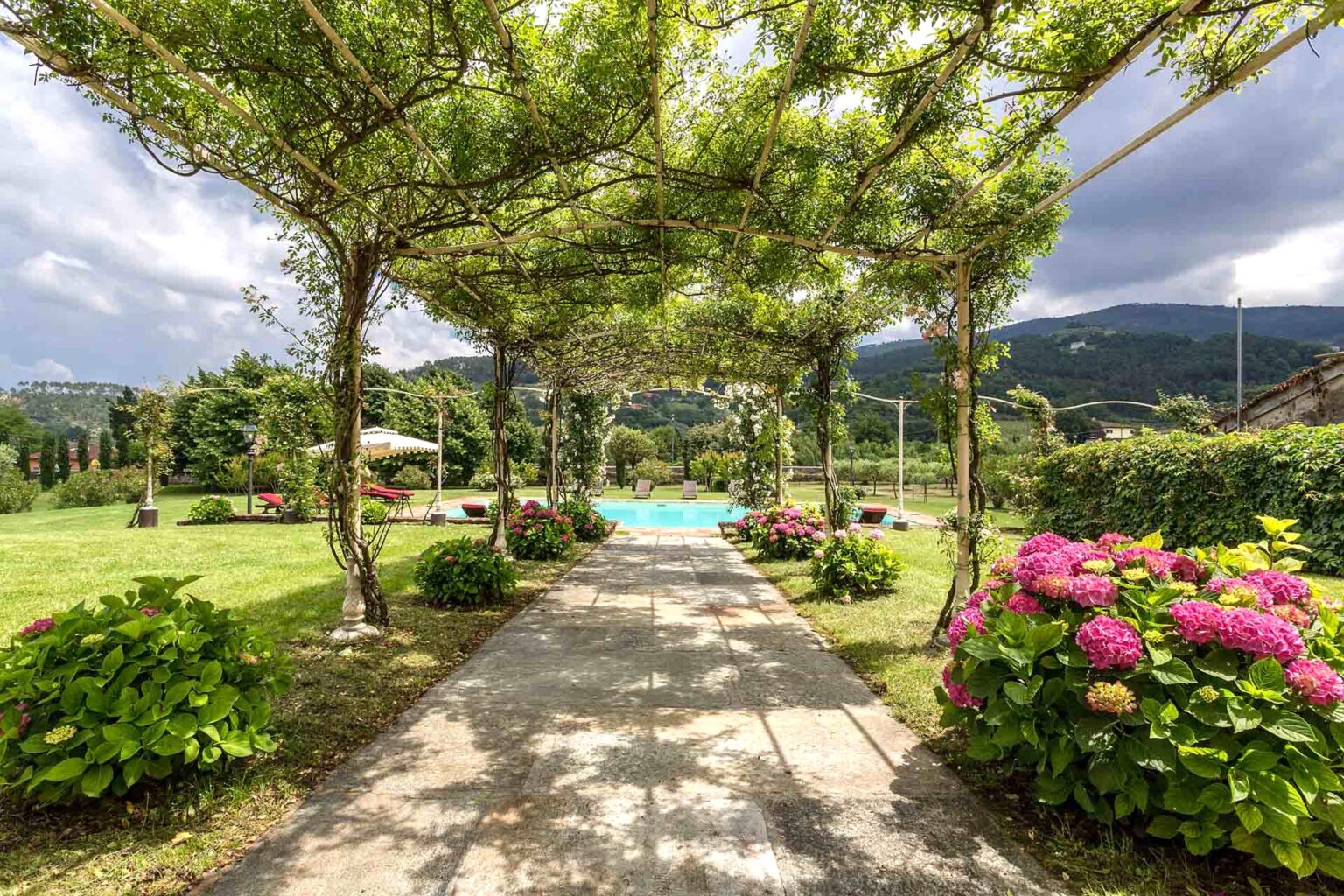 Scenic garden pathway shaded by lush greenery, leading to a sparkling pool surrounded by vibrant flowers and hills in the background. | Only on SopranoVillas