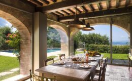 Rustic dining area with a wooden table set for a meal, featuring wine glasses and bottles. Large windows offer a panoramic view of a lush garden and swimming pool, illuminated by natural light and accented by decorative pendant lights.