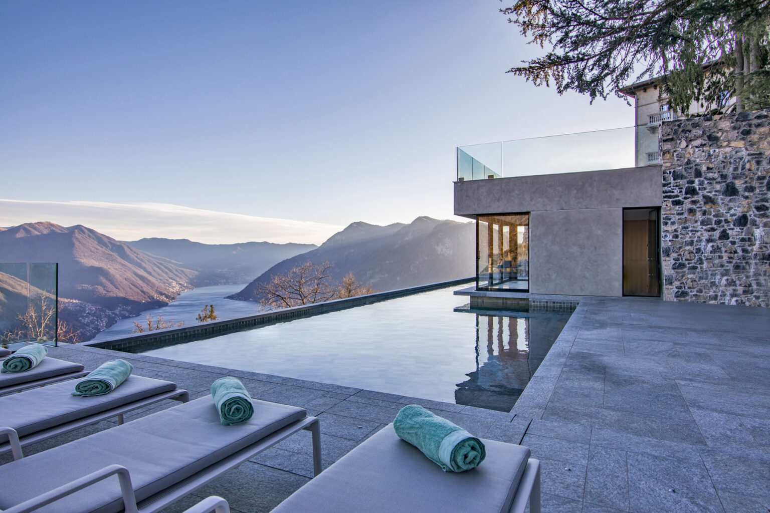 Modern luxury villa with an infinity pool overlooking a mountain landscape at sunset, featuring lounge chairs and a sleek design. | Only on SopranoVillas