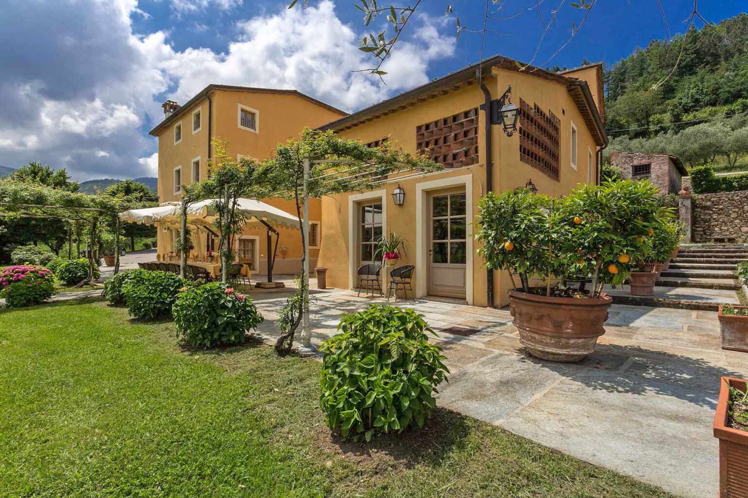 Charming yellow villa surrounded by lush greenery and vibrant flowers, featuring a spacious patio with potted plants and an inviting outdoor seating area, set against a backdrop of rolling hills and a partly cloudy sky. | Only on SopranoVillas