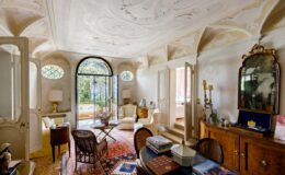 Bright and airy living room featuring elegant vintage decor, ornate ceiling details, large arched windows, and a mix of classic furniture pieces. The space is enhanced by natural light and a cozy atmosphere, showcasing a blend of style and comfort.