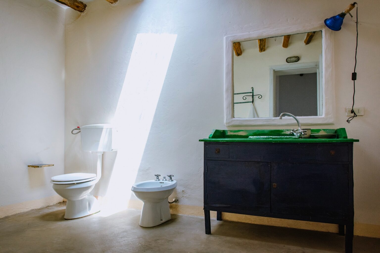 Modern bathroom interior featuring a toilet, bidet, and a stylish dark wood vanity with a green sink. Natural light streams through a large window, enhancing the minimalist design. | Only on SopranoVillas