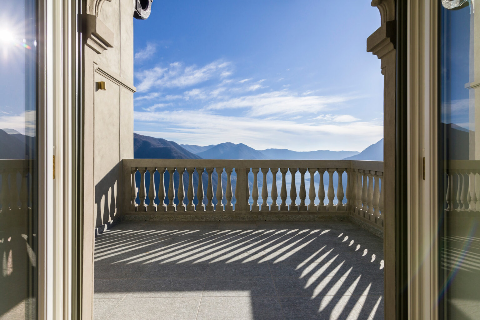 Scenic view from a balcony overlooking mountains and a lake under a clear blue sky, with sunlight casting shadows on the floor. | Only on SopranoVillas