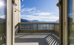 Scenic view from a balcony overlooking mountains and a lake under a clear blue sky, with sunlight casting shadows on the floor.
