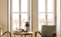 Cozy indoor seating area featuring two chairs and a small table with a vase of flowers, fruit, and a beverage, illuminated by natural light from large windows. Ideal for relaxation and enjoying a view.