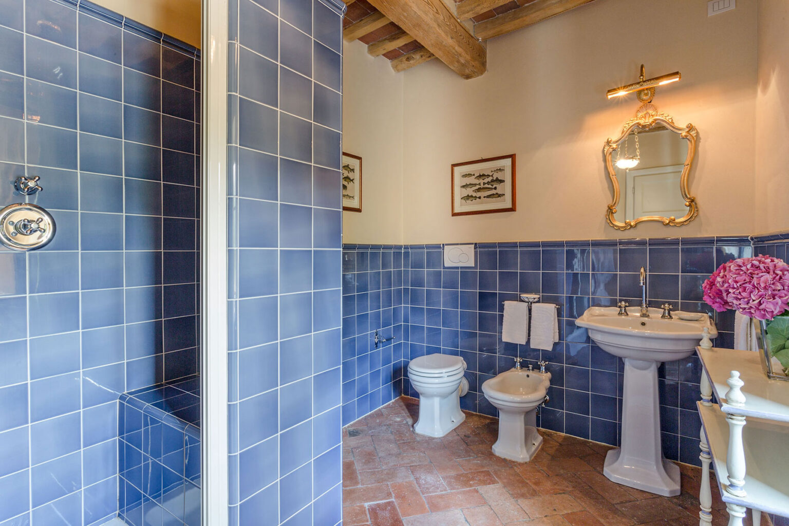 Bright and modern bathroom featuring blue tiled walls, a glass shower, a pedestal sink, a mirror with ornate frame, and decorative accents including fresh flowers. | Only on SopranoVillas