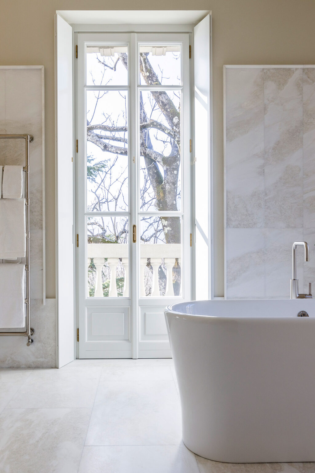 Bright bathroom interior featuring a modern freestanding bathtub, large white double doors opening to a scenic view with trees, and elegant marble walls, creating a serene atmosphere. | Only on SopranoVillas