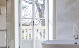 Bright bathroom interior featuring a modern freestanding bathtub, large white double doors opening to a scenic view with trees, and elegant marble walls, creating a serene atmosphere.