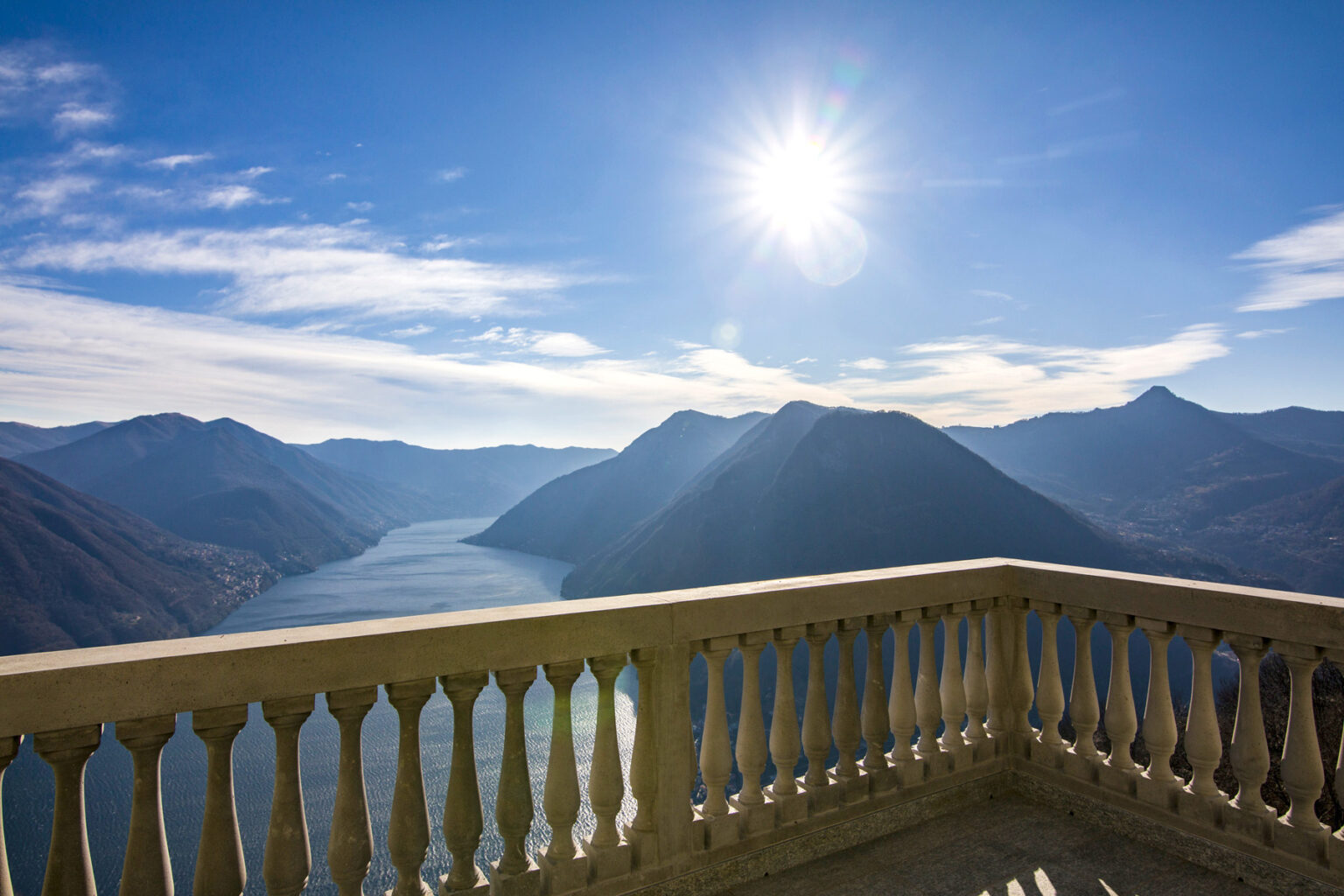 Scenic view from a balcony overlooking a serene lake surrounded by mountains under a bright sun and clear blue sky. | Only on SopranoVillas