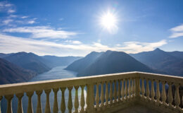 Scenic view from a balcony overlooking a serene lake surrounded by mountains under a bright sun and clear blue sky.