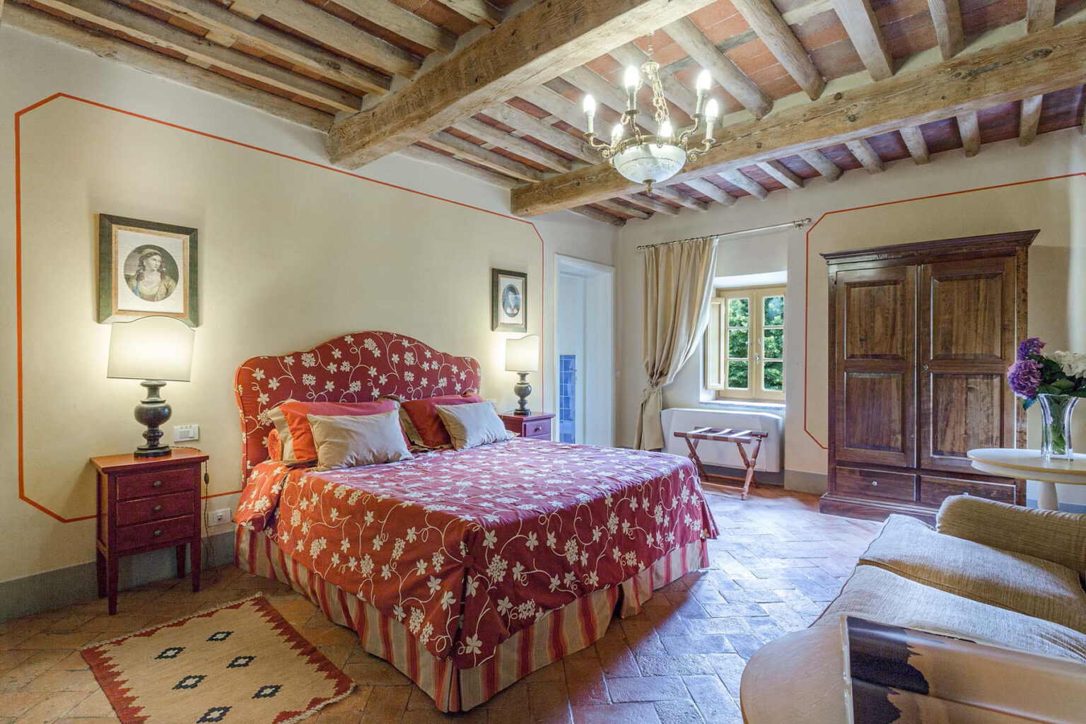 Cozy bedroom featuring a red patterned bedspread, wooden beams on the ceiling, elegant lighting, and decorative artwork. The room includes bedside lamps, a vintage wardrobe, and a window with natural light, creating a warm and inviting atmosphere. | Only on SopranoVillas