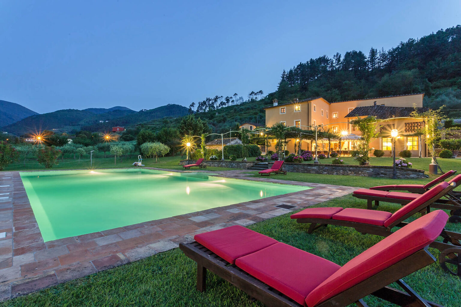 Scenic view of a luxurious villa with a swimming pool at dusk, surrounded by mountains and lush greenery, featuring red lounge chairs and illuminated outdoor spaces. | Only on SopranoVillas