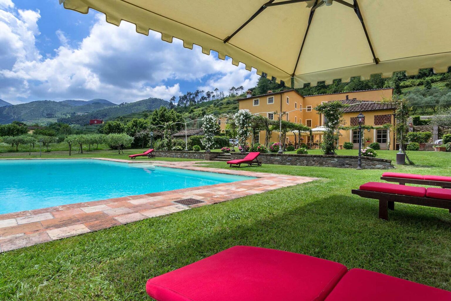 Luxurious outdoor pool area with red lounge chairs under a shaded canopy, surrounded by green grass and picturesque mountains in the background. Ideal for relaxation and leisure in a serene setting. | Only on SopranoVillas