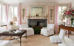 Elegant living room featuring white upholstered furniture, a vintage coffee table, and soft pink curtains. The room is adorned with decorative wall moldings and a central fireplace, complemented by a large mirror and fresh floral arrangements. Natural light floods in through the windows, creating a warm and inviting atmosphere.