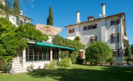 Elegant villa surrounded by lush greenery, featuring a spacious garden, outdoor seating area with an umbrella, and classic architectural details under a clear blue sky.
