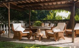Cozy outdoor seating area under a wooden pergola, featuring wicker chairs with white cushions and a small table, surrounded by greenery. Ideal for relaxation and entertaining guests.