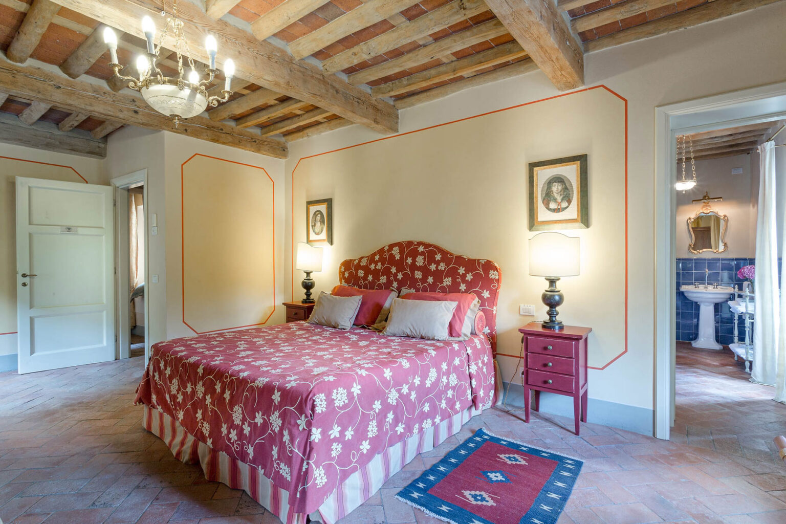 Cozy bedroom featuring a red patterned bedspread, vintage decor, and wooden beams. The room includes bedside lamps, framed portraits on the walls, and an adjoining bathroom visible through the open door. Ideal for a charming and relaxing stay. | Only on SopranoVillas