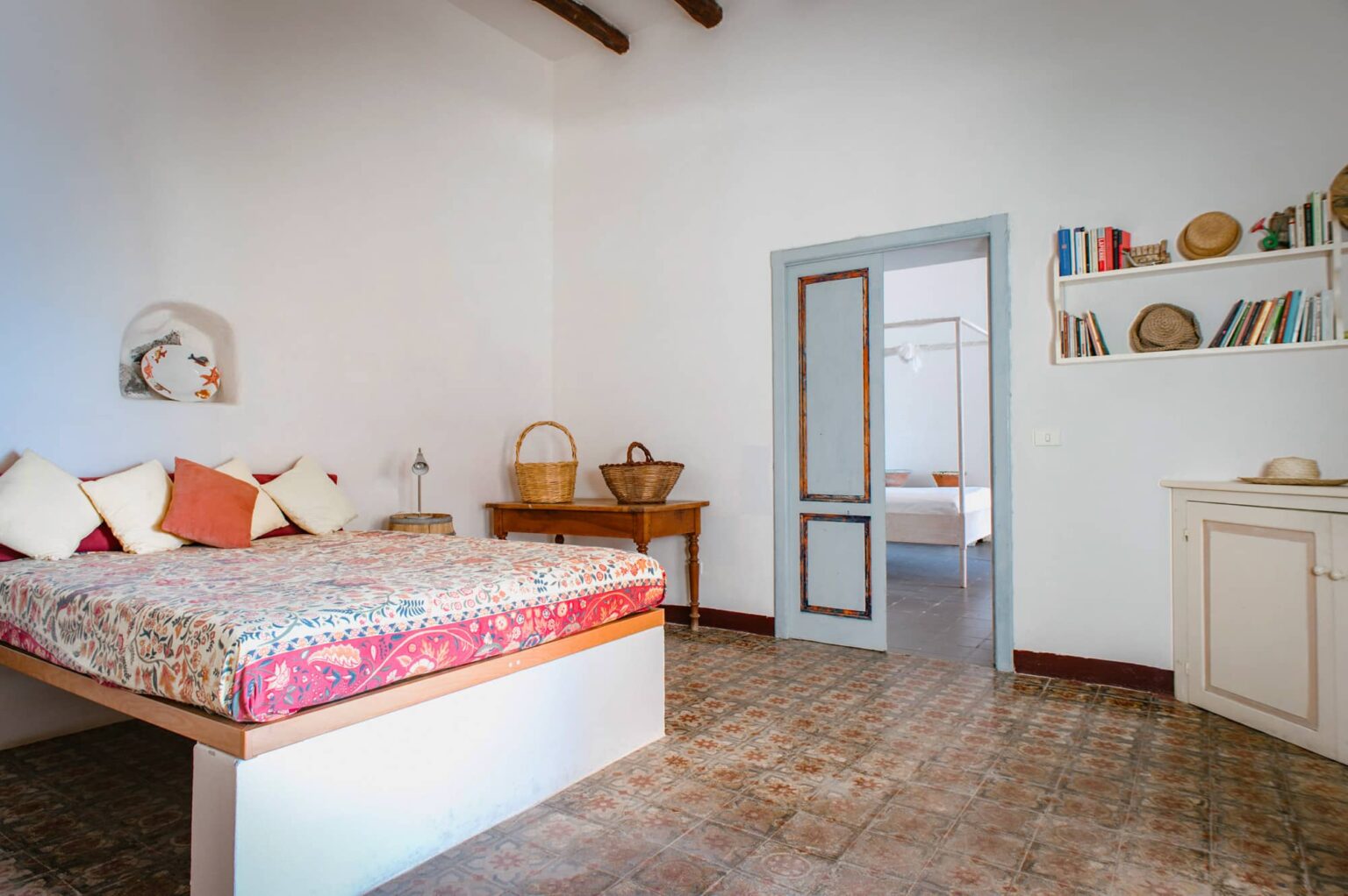 Cozy and bright bedroom featuring a large bed with colorful bedding, wooden furniture, and a door leading to another room. The space showcases a minimalist design with warm tones and natural light, ideal for relaxation. | Only on SopranoVillas