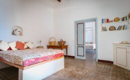 Cozy and bright bedroom featuring a large bed with colorful bedding, wooden furniture, and a door leading to another room. The space showcases a minimalist design with warm tones and natural light, ideal for relaxation.