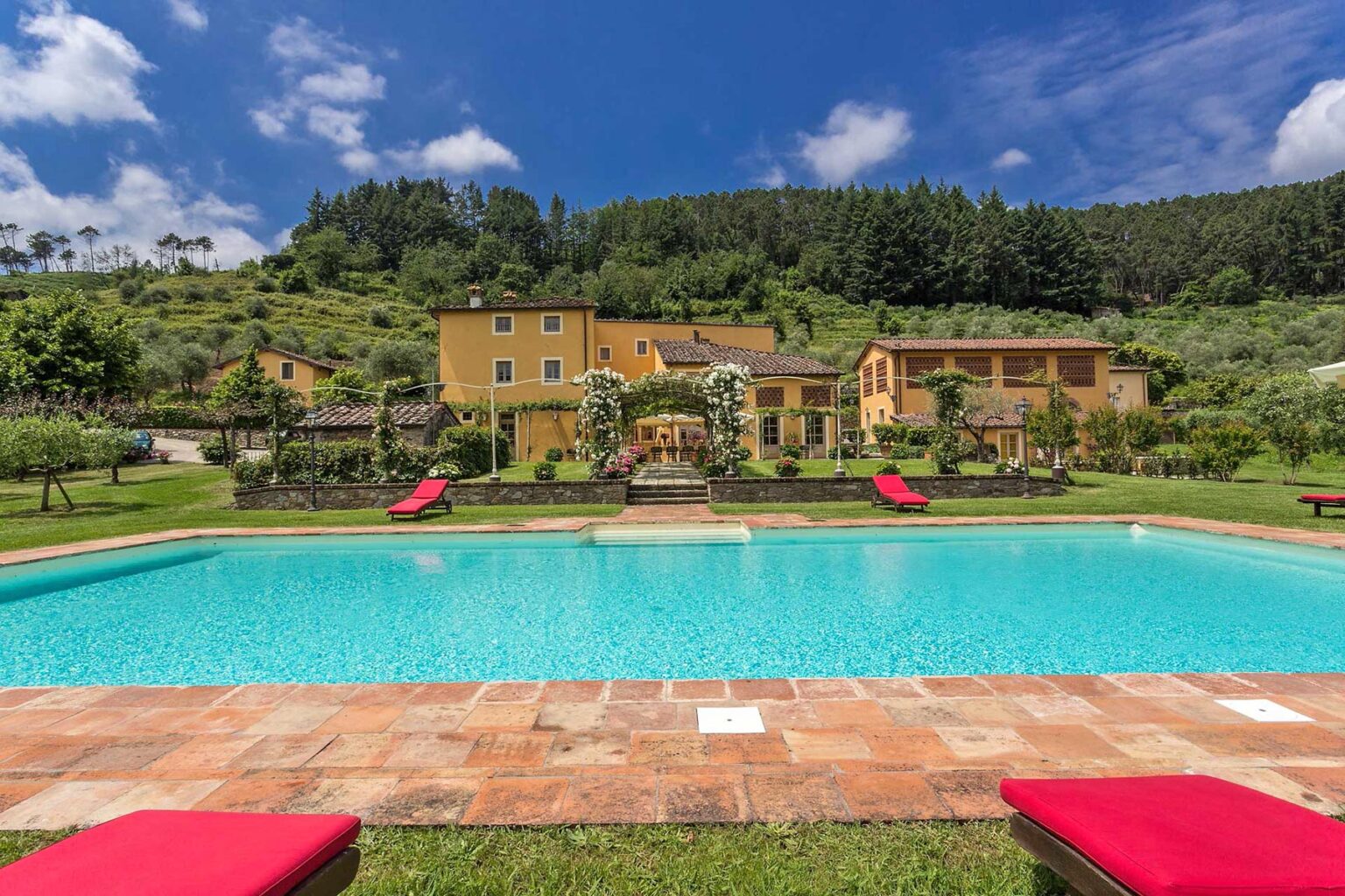Scenic view of a luxurious villa with a swimming pool, surrounded by lush greenery and hills, featuring red lounge chairs and a sunny sky. Ideal for vacation rentals and outdoor relaxation. | Only on SopranoVillas