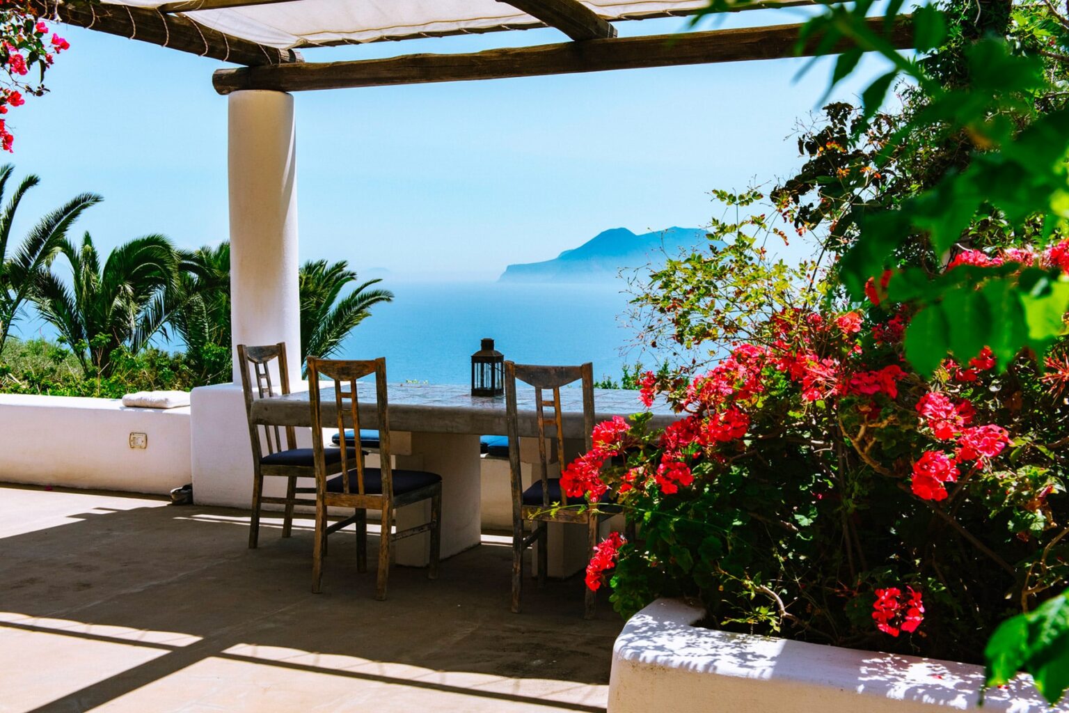 Cozy outdoor seating area with wooden chairs and a table, surrounded by vibrant flowers, overlooking a serene ocean view and distant mountains. Perfect for relaxation and enjoying nature. | Only on SopranoVillas