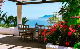 Cozy outdoor seating area with wooden chairs and a table, surrounded by vibrant flowers, overlooking a serene ocean view and distant mountains. Perfect for relaxation and enjoying nature.