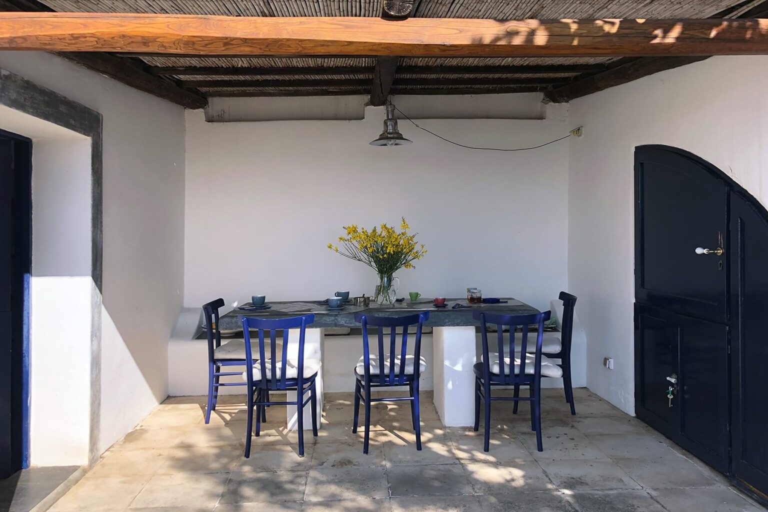 Outdoor dining area with blue chairs and a table set for a meal, featuring a vase of yellow flowers, under a rustic wooden roof. Ideal for gatherings in a serene environment. | Only on SopranoVillas
