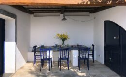 Outdoor dining area with blue chairs and a table set for a meal, featuring a vase of yellow flowers, under a rustic wooden roof. Ideal for gatherings in a serene environment.
