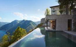 Modern luxury villa with an infinity pool overlooking a scenic mountain landscape and a tranquil lake under a clear blue sky.