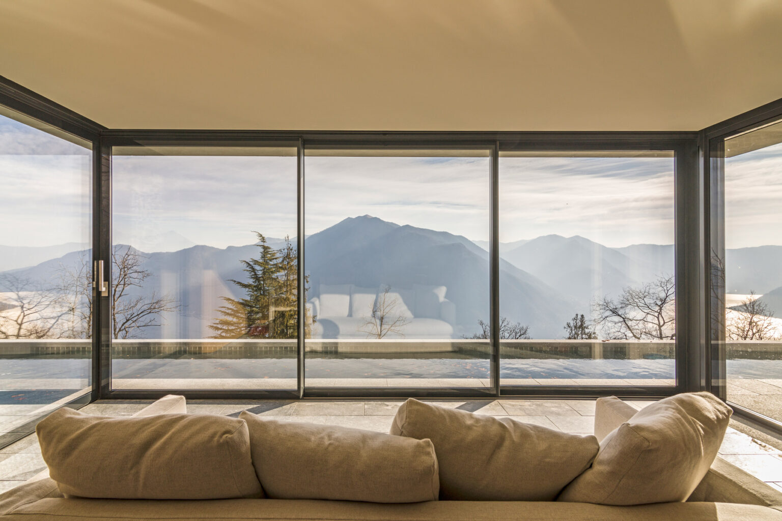 Modern living room with large glass windows showcasing a panoramic view of mountains and trees. A cozy beige sofa is positioned in front, creating a serene and inviting atmosphere. | Only on SopranoVillas
