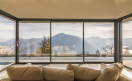 Modern living room with large glass windows showcasing a panoramic view of mountains and trees. A cozy beige sofa is positioned in front, creating a serene and inviting atmosphere.
