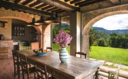Rustic dining area with a wooden table and chairs, featuring a vase of purple flowers, set in an open space with arched windows overlooking a green landscape and mountains. Ideal for outdoor dining and entertaining.