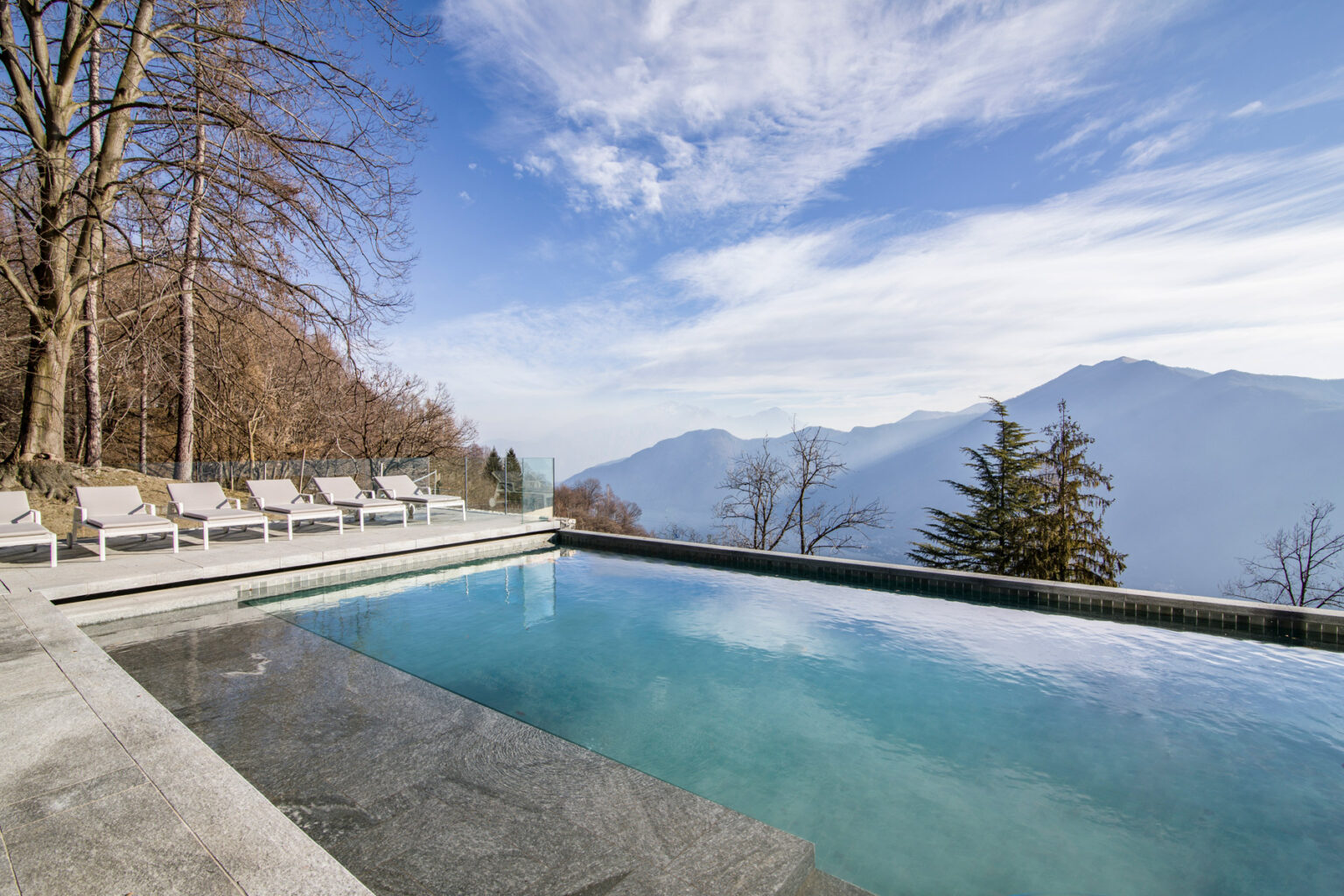 Modern infinity pool with mountain views, surrounded by lounge chairs and trees, under a clear blue sky. Ideal for luxury relaxation and outdoor living. | Only on SopranoVillas
