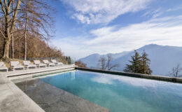 Modern infinity pool with mountain views, surrounded by lounge chairs and trees, under a clear blue sky. Ideal for luxury relaxation and outdoor living.