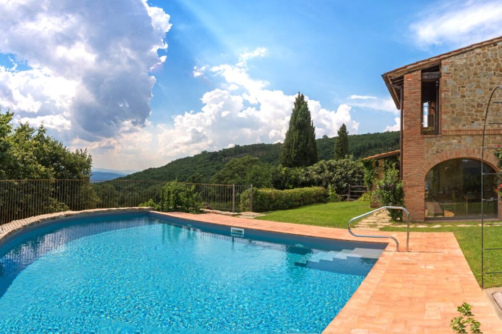 Scenic view of a sparkling blue swimming pool surrounded by lush greenery and a rustic stone building under a bright sky, ideal for vacation rentals in the countryside. | Only on SopranoVillas
