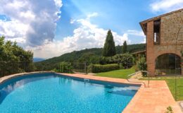 Scenic view of a sparkling blue swimming pool surrounded by lush greenery and a rustic stone building under a bright sky, ideal for vacation rentals in the countryside.