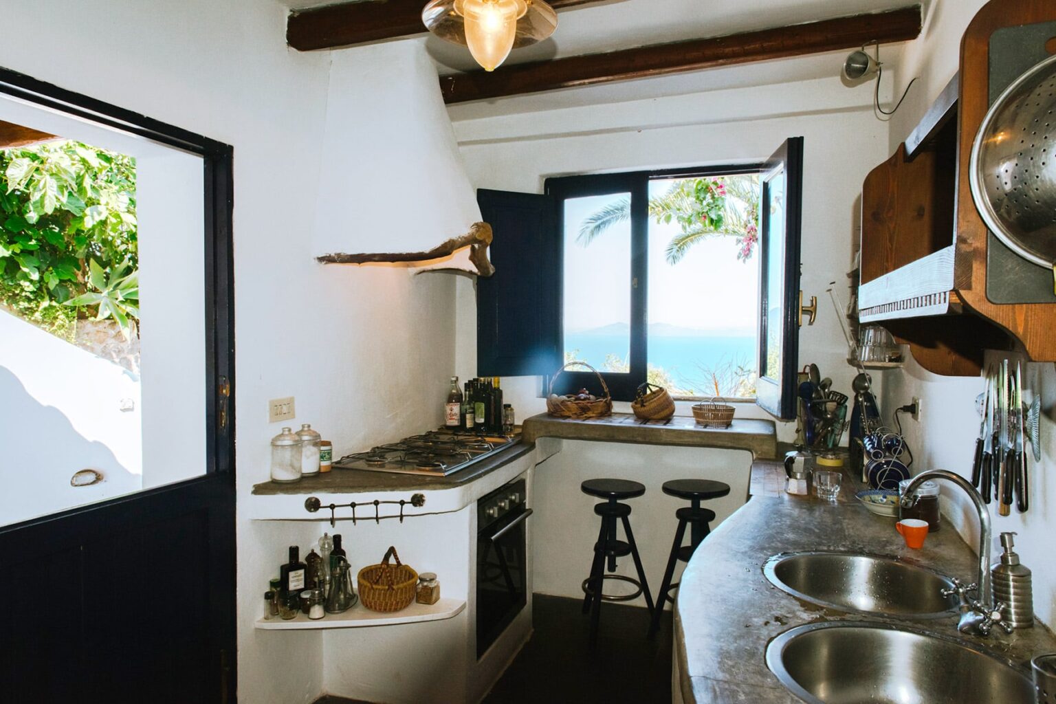 Cozy kitchen with a coastal view, featuring a modern gas stove, dual sinks, wooden shelves, and bar stools, surrounded by white walls and wooden beams. | Only on SopranoVillas