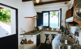 Cozy kitchen with a coastal view, featuring a modern gas stove, dual sinks, wooden shelves, and bar stools, surrounded by white walls and wooden beams.